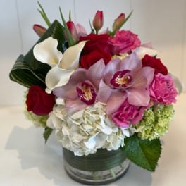 Compact arrangement of white hydrangeas, red roses, pink orchids, tulips, and calla lilies in a glass vase.