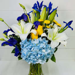 Tall arrangement of white lilies, blue hydrangeas and irises, and yellow roses in a clear glass vase