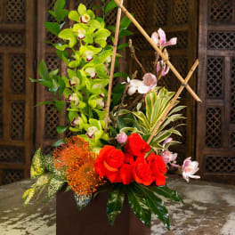 Tall floral arrangement with red roses, green orchids, and orange blooms in a square vase