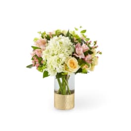 Pastel bouquet of white hydrangeas, yellow roses, and pink blooms in a clear vase with a gold base.