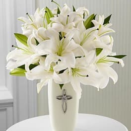 White lilies arranged in a tall cream vase with a silver cross emblem.