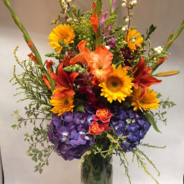 Tall mixed bouquet with sunflowers, lilies, and purple hydrangeas in a glass vase