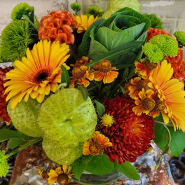 Bouquet of yellow gerbera daisies, orange mums, and green ornamental pods in a glass vase