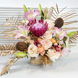 Modern arrangement with pink lilies, roses, a central protea and gold-painted leaves in a square glass vase