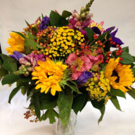 Mixed bouquet of sunflowers, pink blooms, and yellow filler flowers in a glass vase