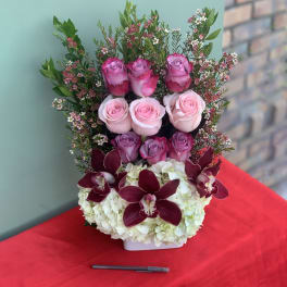 Tiered floral arrangement with pink roses, white hydrangeas, and dark orchids in a white container