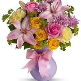 Pink and yellow mixed bouquet in a lavender vase with a pink ribbon