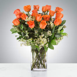 Tall arrangement of orange roses with small white blooms in a clear rectangular glass vase.