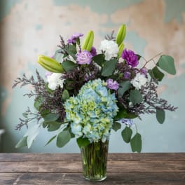 Mixed bouquet with blue hydrangea, purple and white blooms, and lily buds in a clear glass vase.