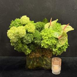 Green floral arrangement in a glass vase with an anthurium and candle