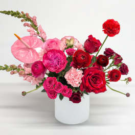 Red and pink roses, ranunculus, and mixed blooms arranged in a white cylinder vase.