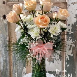 Peach roses and white daisies in a tall green glass vase with a pink bow