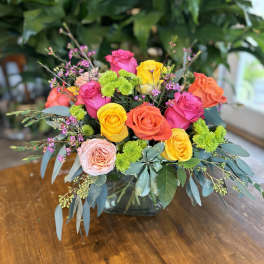 Bright arrangement of pink, orange and yellow roses with green accents in a clear glass cube vase.