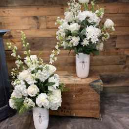Two white floral arrangements in tall white vases on a wooden surface