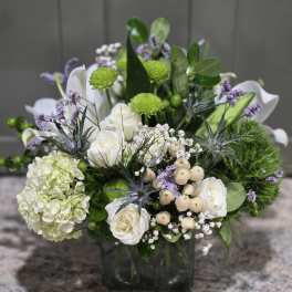 Low arrangement of white roses, hydrangea, green mums, and lavender accents in a glass cube vase