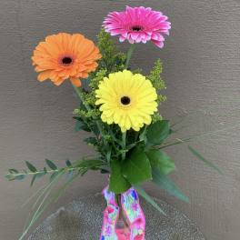 Three colorful gerbera daisies in a glass vase with a floral ribbon