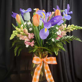 Vase of purple irises and orange tulips with pink blooms and a gingham bow.