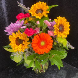 Bright mixed bouquet with sunflowers, orange gerbera, carnations, and mums in a clear glass vase