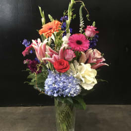 Tall mixed bouquet in a clear glass vase with pink, orange, blue, and white flowers.