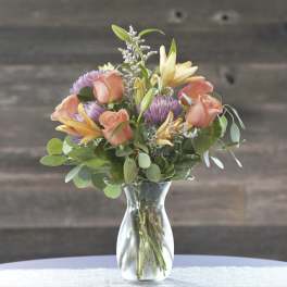 Mixed bouquet of peach roses, lilies, and purple blooms in a clear glass vase