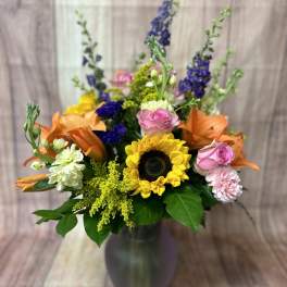 Mixed bouquet in a tall vase with sunflowers, roses, lilies, and purple spikes