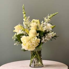 Arrangement of soft yellow roses, lilies, and hydrangeas in a clear glass vase on a round table.