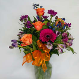 Mixed bouquet of orange roses, purple flowers, and a pink gerbera in a glass vase