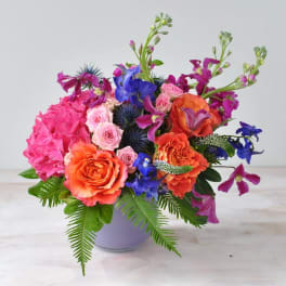 Bright mixed bouquet in a lavender vase with pink, orange, and blue flowers
