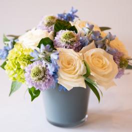 Bouquet of cream roses, blue flowers, and lavender blooms in a gray vase
