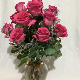 Arrangement of bright pink roses with filler blooms in a clear glass vase tied with a gold ribbon.