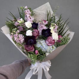 Hand-tied bouquet of pink, white, and dark purple flowers wrapped in paper