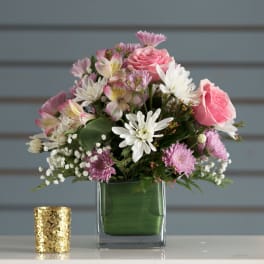 Compact pink and white mixed flower arrangement in a square glass vase with a gold votive holder beside it