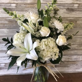 White floral arrangement in a glass vase with lilies, roses, and hydrangeas