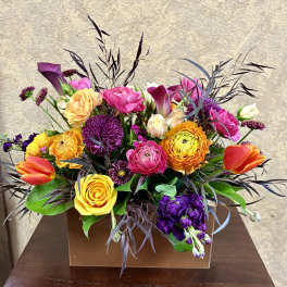 Low rectangular arrangement of bright yellow, orange, pink, and purple flowers in a brown box.