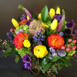 Bright mixed arrangement of yellow tulips, orange roses and purple blooms in a clear glass vase