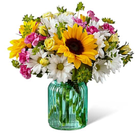 Bouquet of sunflowers, white daisies, pink carnations, and yellow roses in a green glass vase