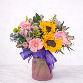 Mixed bouquet of sunflowers, pink daisies, and purple flowers in a vase with a purple ribbon