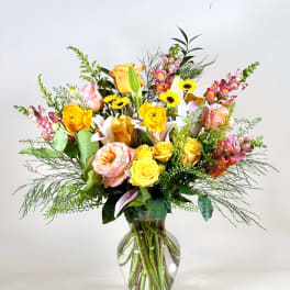 Tall mixed bouquet of yellow and peach roses, lilies, daisies, and snapdragons in a clear glass vase