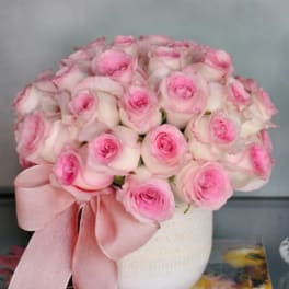 Pink and white roses arranged in a white vase with a pink ribbon