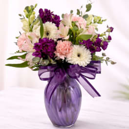 Mixed bouquet of pink, white, and purple flowers in a purple glass vase with a matching bow