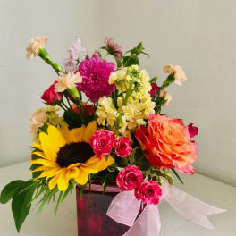 Bright mixed arrangement with sunflower, roses, and carnations in a pink cube vase with ribbon