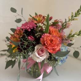 Mixed bouquet of pink, orange, and blue flowers in a glass vase with a pink ribbon