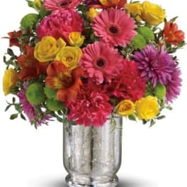 Bright mixed bouquet of pink gerbera daisies, yellow roses, and green buttons in a silver glass vase