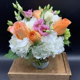 Short arrangement of peach roses, white hydrangeas, and pink lisianthus in a clear glass vase.