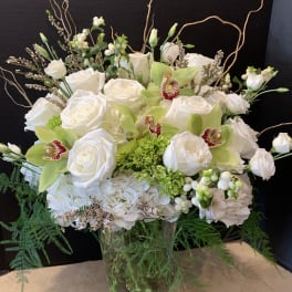 Tall white and green arrangement with roses, hydrangea, and cymbidium orchids in a clear glass vase