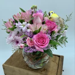 Round glass vase of pink and lavender flowers with a small yellow bird pick on a wooden box