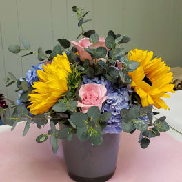 Arrangement of yellow sunflowers, pink roses, and blue hydrangeas in a gray vase