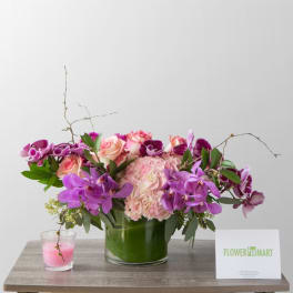 Pink and purple floral arrangement in a glass vase with a candle and card