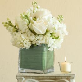 White floral arrangement in a square glass vase beside a lit candle