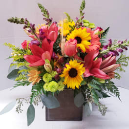 Bright mixed arrangement with red lilies, yellow sunflowers, and roses in a square wooden vase